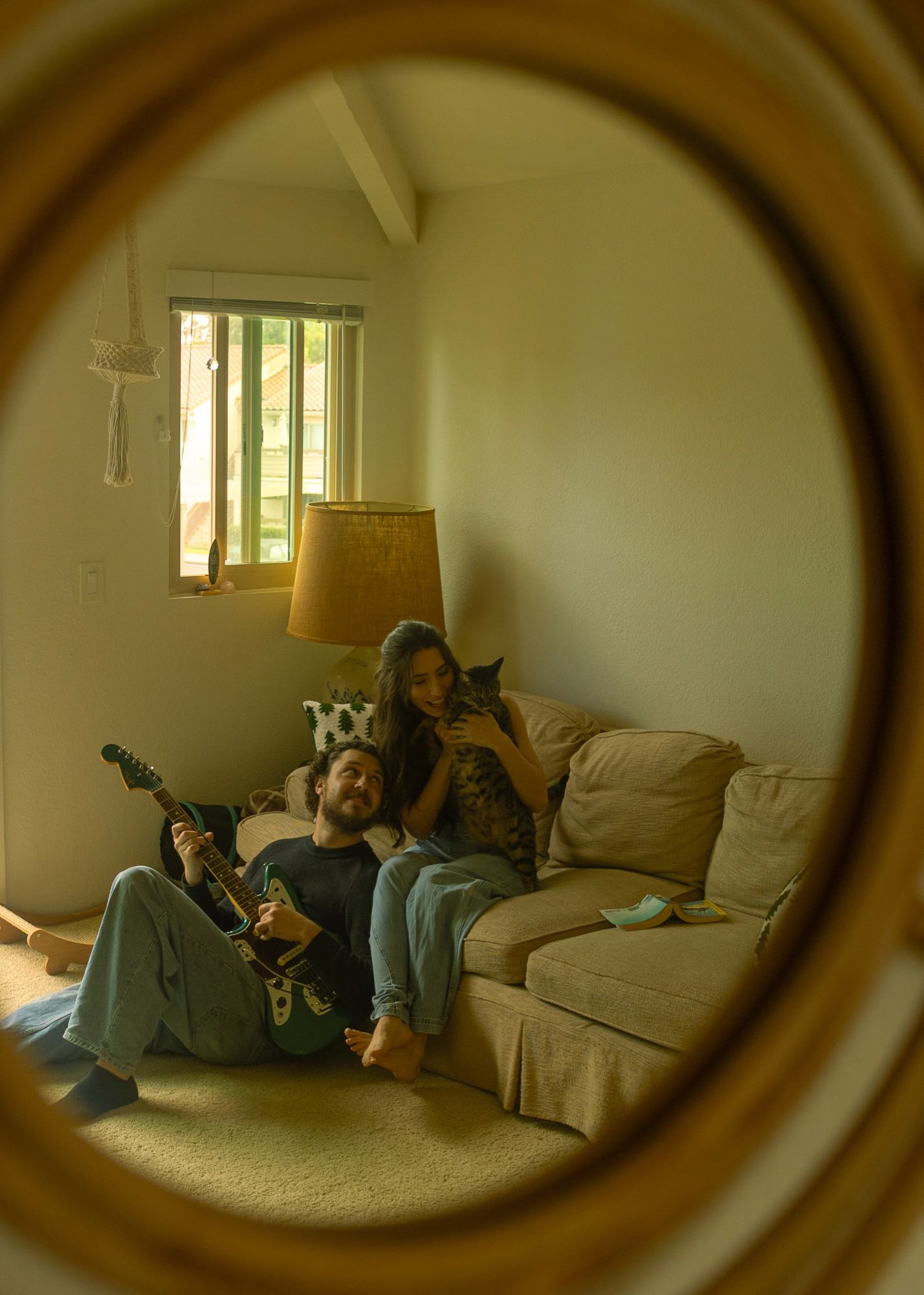 Jordan with a guitar and Katherine holding Mango the cat on the couch, seen in a round mirror