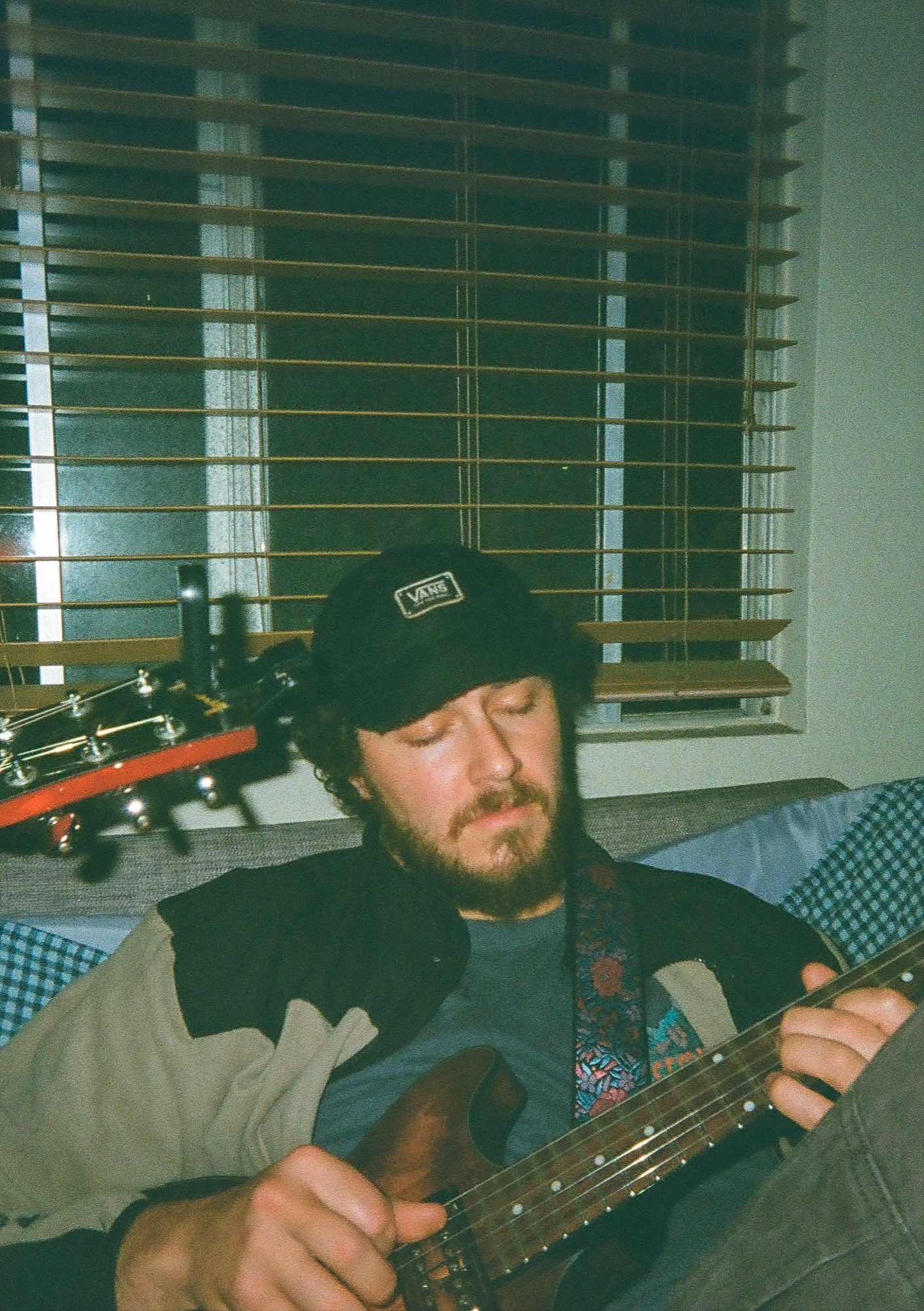 Jordan in a cap, eyes down, playing guitar on a couch in front of a window with the blinds down