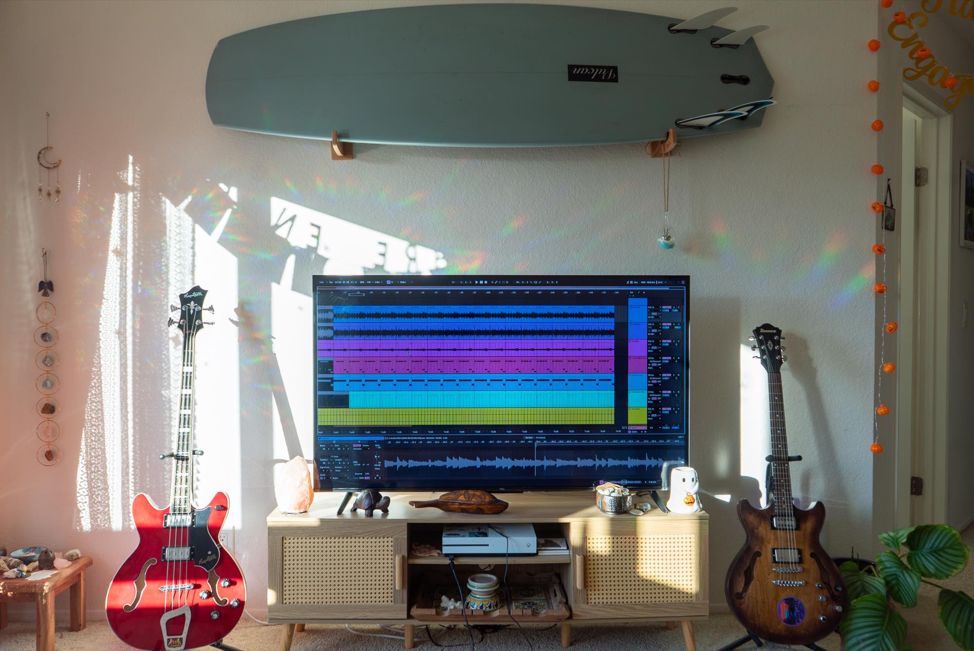 A red electric guitar and a dark bass guitar beside the studio desk, with a surfboard mounted above